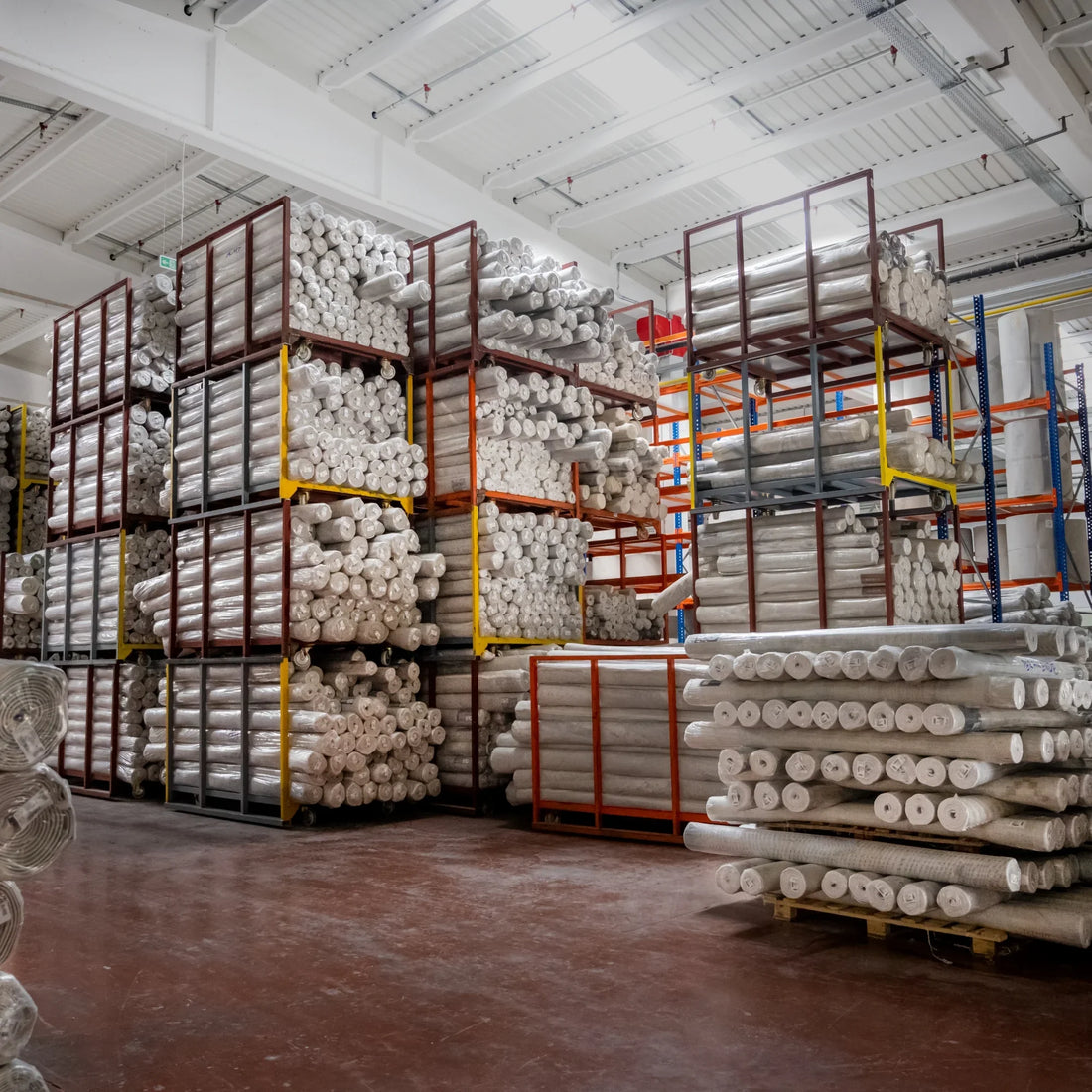 Büyük bir depo içinde istiflenmiş, çeşitli boy ve desenlerde rulo halinde paketlenmiş halılar