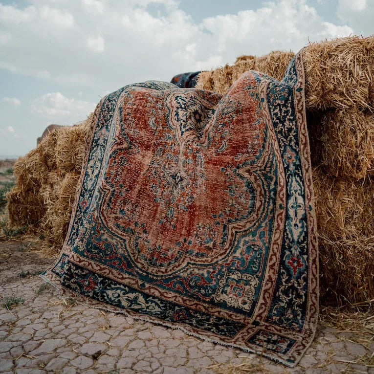 Açık havada balya üzerine serilmiş vintage desenli, eskitme halı; kırmızı ve mavi klasik motifli.