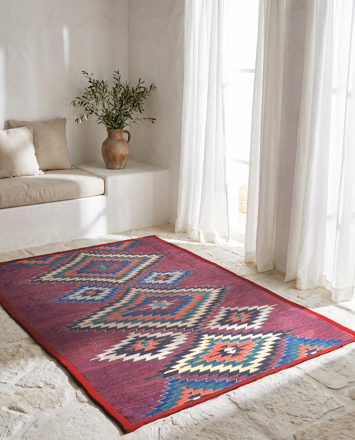 Kireç badanalı Ege tarzı odada taş zemin üzerinde bordo zeminli kilim desenli halı